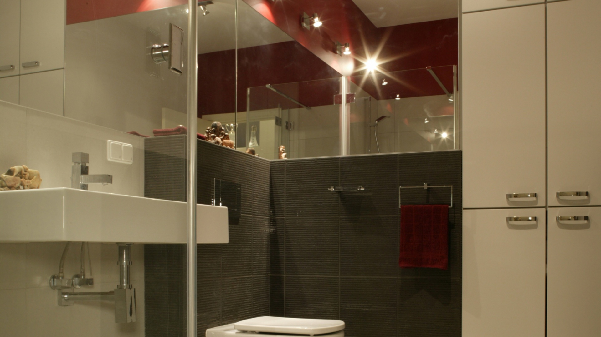 Ciemne płytki (Marazzi) pokrywające fragment ściany w okolicy sedesu mają charakterystyczną, żłobioną fakturę. Czerwień nad lustrem, w połączeniu z nowoczesnym oświetleniem, ożywia i rozjaśnia wnętrze. Fot. Monika Filipiuk.