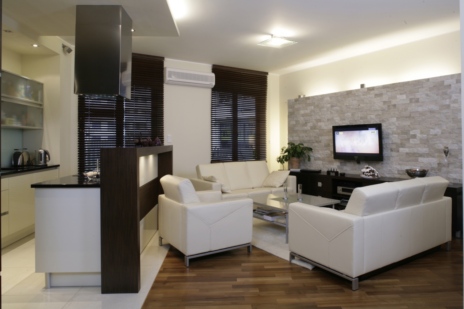 Zarówno salon, jak i kuchnia zostały urządzone w tym samym minimalistycznym stylu – tylko niezbędne sprzęty w prostych, eleganckich liniach. Ciepłą nutę wprowadza podłoga pokryta akacją parzoną i przesłaniające okna drewniane żaluzje. Fot. Monika Filipiuk.