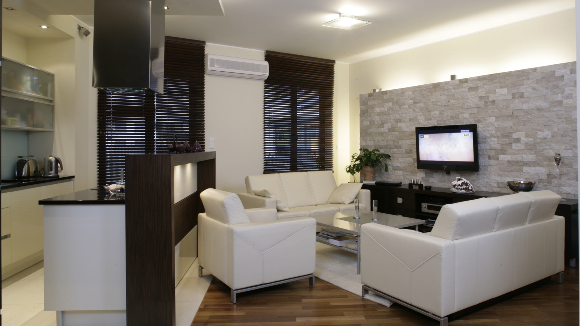 Zarówno salon, jak i kuchnia zostały urządzone w tym samym minimalistycznym stylu – tylko niezbędne sprzęty w prostych, eleganckich liniach. Ciepłą nutę wprowadza podłoga pokryta akacją parzoną i przesłaniające okna drewniane żaluzje. Fot. Monika Filipiuk.