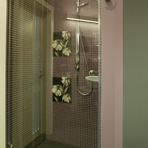 W przesłoniętej szklaną taflą kabinie prysznicowej, obok fioletowej mozaiki na ścianie (Marazzi), pojawiły się kwieciste kafle (Dado Ceramica) oraz pionowe wstawki ze szkła, wpuszczające do wnętrza dzienne światło. Fot. Bartosz Jarosz.
