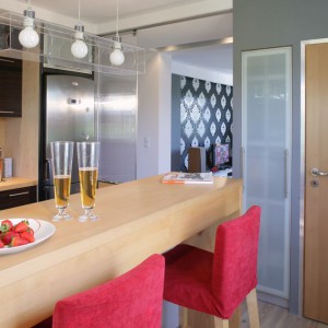 Przy kuchennym barze z drewna jesionowego ustawiono parę krzeseł barowych. Krzesła pochodzą z IKEA, ale ich indywidualne czerwone obicie nadała im architekt, dzięki czemu stały się bardziej wyraziste i odważne. Strefa ta ma własne oświetlenie w postaci designerskiej lampy z transparentnego pleksi (Trends). Fot. Monika Filipiuk.