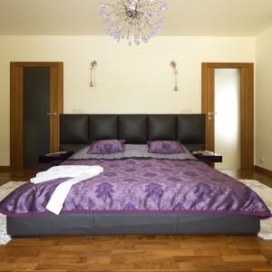 Duże, tapicerowane łoże jest głównym meblem w sypialni. Zdobi je fioletowa połyskująca narzuta z klasycznym ornamentem. Do niej dobrano kryształowy żyrandol w lawendowym odcieniu. Projekt Tomasz Tubisz Fot. Przemysław Andruk