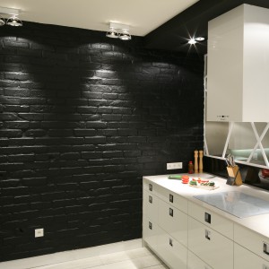 Najmocniejszym elementem kuchni jest ceglana ściana pomalowana czarną farbą ceramiczną. Stanowi ciekawy kontrast dla białych mebli w połysku. Projekt: Dominik Respondek. Fot. Bartosz Jarosz. 