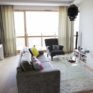Przytulna, miękka sofa, stylizowany tapicerowany fotel i miękki dywan budują w salonie przytulny klimat. Na ściance działowej oddzielającej salon od sypialni zawieszono telewizor. Szafka RTV komponuje się kolorystycznie ze ścianą. Z czernią ekranu harmonizuje czarny, dekoracyjny żyrandol. Projekt: Chalupko Design. Fot. Chalupko Design.