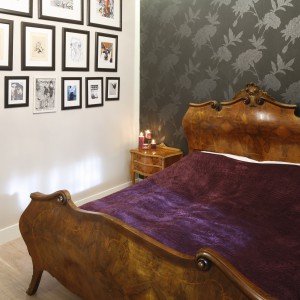 Wygodne łoże z pięknym, drewnianym zagłówkiem zajmuje główne miejsce w sypialni. Doskonale prezentuje się na tle ściany pokrytej ciemną tapetą w delikatne wzory. Projekt: Monika Gorlikowska. Fot. Bartosz Jarosz.