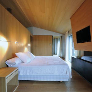 Unikalna sypialnia master zachwyca ilością użytego w niej drewna. Materiał znajduje się na suficie, zabudowie nad kominkiem oraz na ściance, przy której stanęło podwójne łóżko. Projekt: Coblonal Arquitectura. Fot. Coblonal Arquitectura.