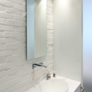  Surowa powierzchnia cegły malowanej białą farbą wprowadza do łazienki loftowy klimat, a  jednocześnie świetnie komponuje się z elegancką powierzchnią lakierowanych frontów meblowych. Fot. Bartosz Jarosz.