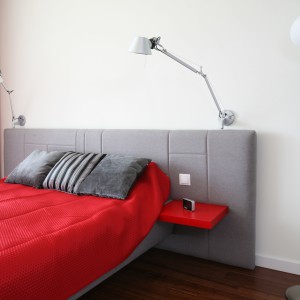Szary tapicerowany zagłówek jest dokładnie w takim samym kolorze jak rama łóżka. Doskonale pasuje zarówno do jasnej ściany, jak i czerwonych akcentów ożywiających sypialnie. Projekt: Iza Szewc. Fot. Bartosz Jarosz.