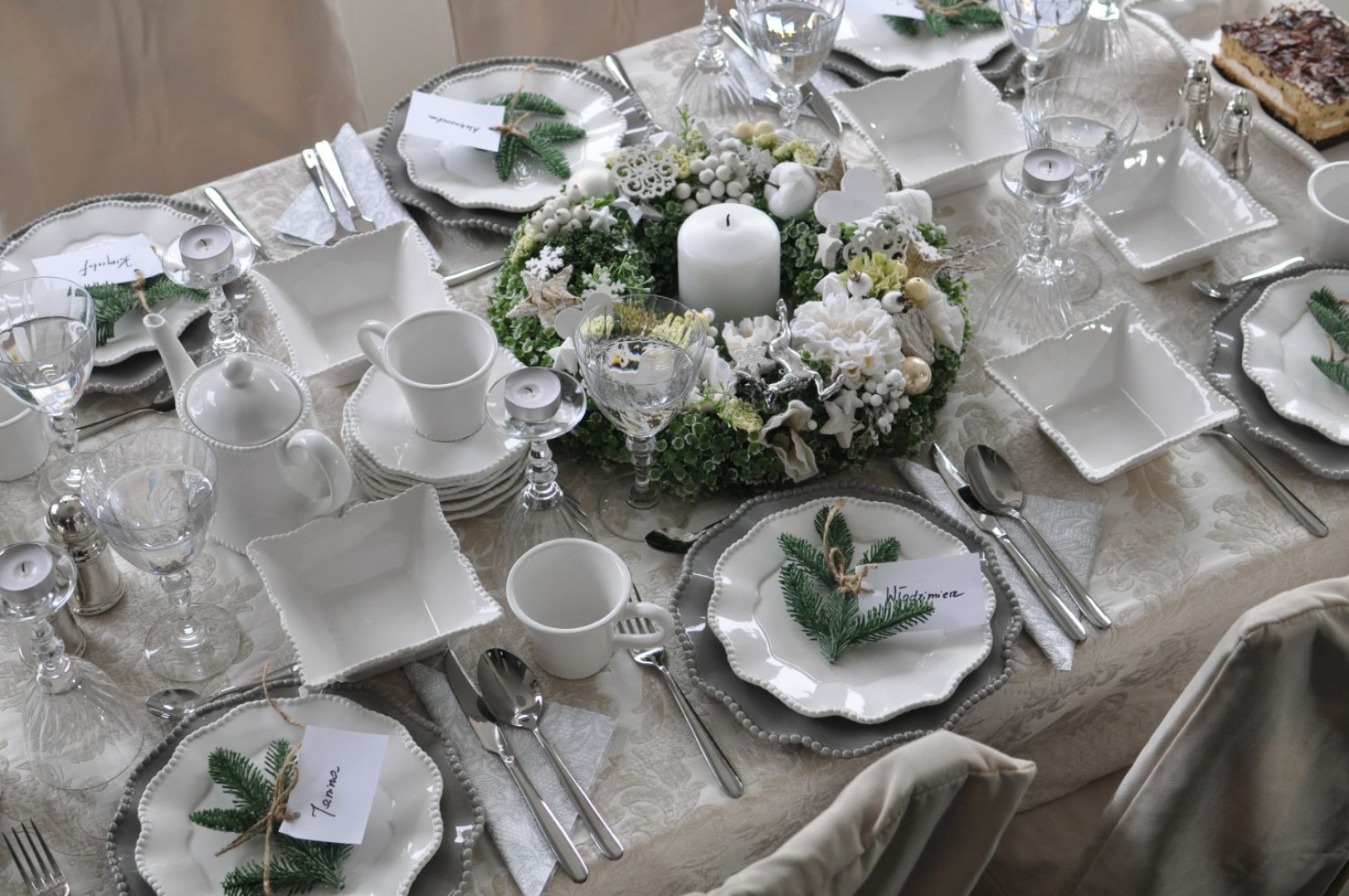 Elegancki obrus w jasnobeżowym kolorze zdobią subtelne, wyszywane wzory. Z kolorystyką obrusu harmonizują duże talerze. Oprócz tych dwóch elementów na stole króluje biel i świąteczne, zielone gałązki świerku. W środku aranżacji ułożono piękny, ręcznie robiony wianek. Fot. Tendom.pl.