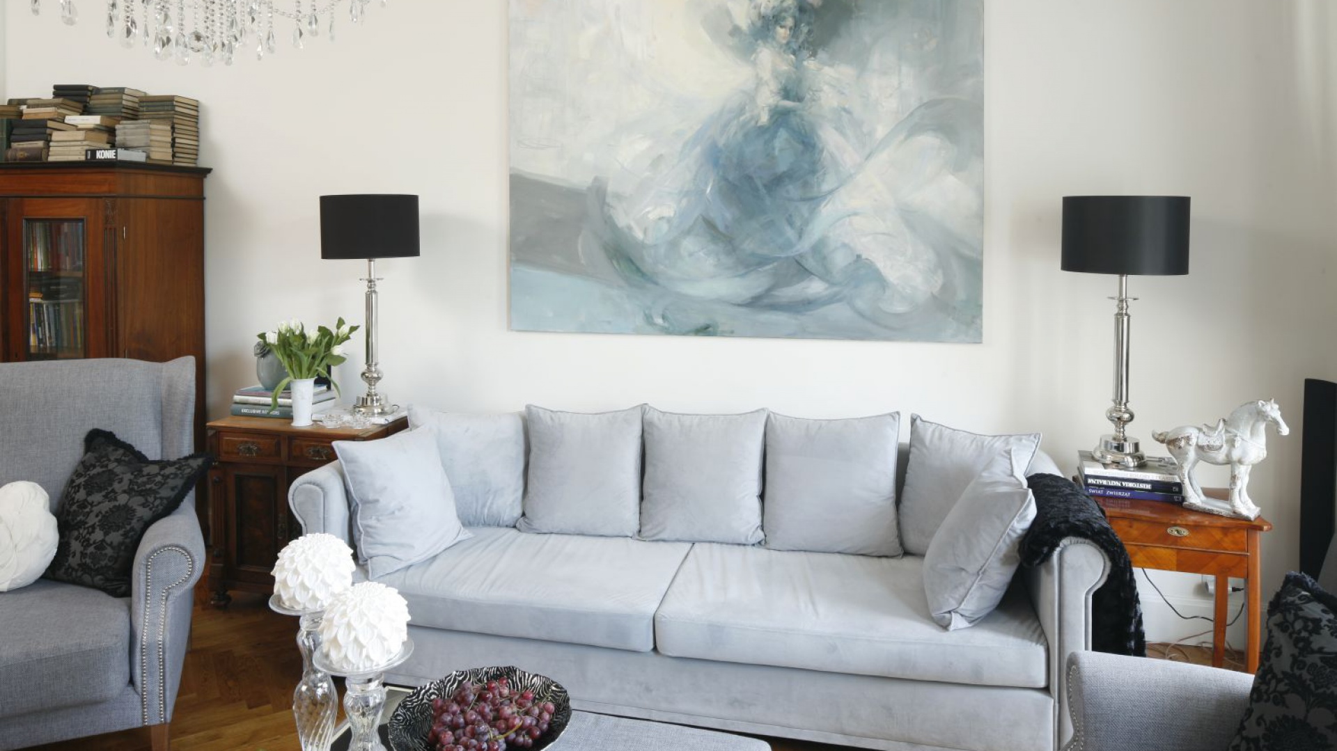 Stary drewniany regał oraz stylizowane komódki po obu stronach kanapy wpisują się w klasyczną aranżację salonu i idealnie współgrają z eleganckimi lampami na stylizowanych nogach. Projekt: Iwona Kurkowska. Fot. Bartosz Jarosz