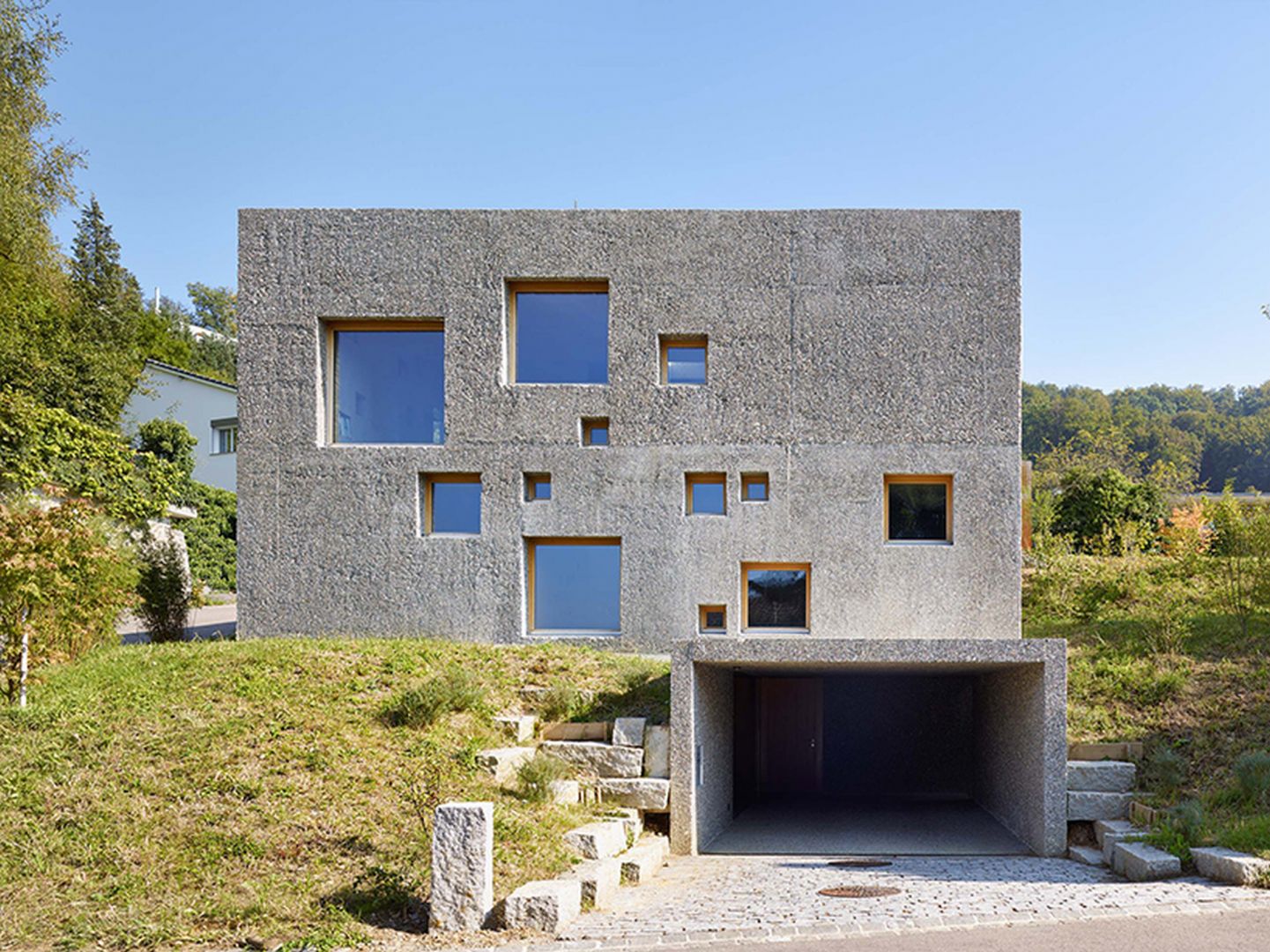 Ta betonowa 2-piętrowa „kostka” znajduje się w północnej Szwajcarii w dzielnicy Füllinsdorf. Rezydencja z oryginalną, betonową elewacją, wyróżnia się finezyjnie rozmieszczonymi, małymi i dużymi kwadratowymi oknami. Fot. Hannes Henz