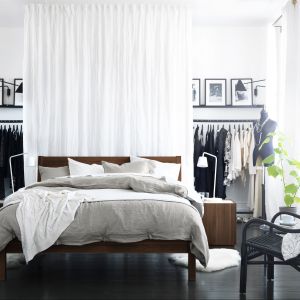 Łóżko wykonane w ciemnym drewnie idealnie współgra z czarno białym pomieszczeniem, zaś garderoba to wieszaki za zasłoną. Fot. IKEA
