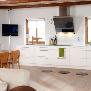 Białe meble kuchenne pięknie prezentują się w towarzystwie beżowej podłogi, drewnianego stołu i wyeksponowanych belek stropowych. Drewnianą podłogę ozdabiają szare, dekoracyjne chodniczki. Fot. Ballin 