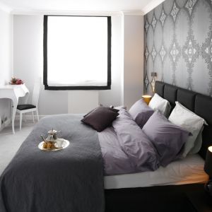 Łóżko z miękkim, tapicerowanym zagłówkiem to również najprostszy sposób na wizualne ocieplenie sypialni. Ustawione na tle szarej, dekoracyjnej tapety będzie zdobiło sypialnię. Projekt: Magdalena Smyk. Fot. Bartosz Jarosz