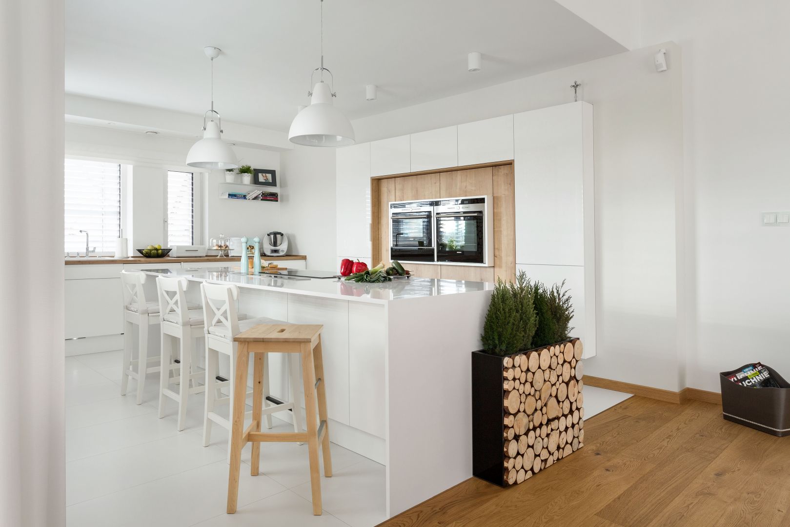 Biała kuchnia w minimalistycznym stylu sprawi, że wnętrze będzie świetliste i łagodne w odbiorze. W tym wnętrzu wyspa pełni rolę miejsca gotowania i przygotowywania posiłków. Umieszczono w niej okap wysuwany z blatu. Projekt: Małgorzata Błaszczak. Fot. Artur Krupa