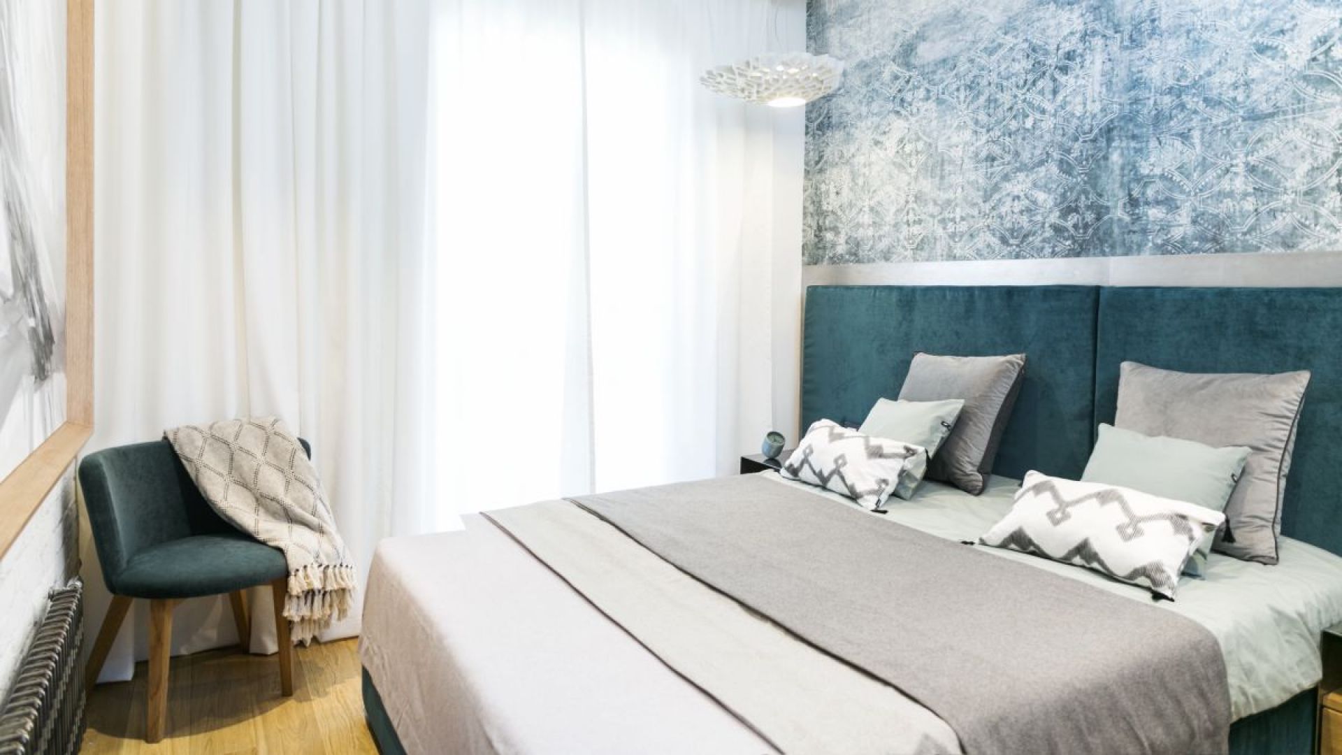 W sypialni, w której dominują biel i butelkowa zieleń, postawiono na welurową tkaninę o zdecydowanym, głębokim kolorze. Tapicerowane łóżko zwieńczono wezgłowiem ujętym w ramę z patynowanej blachy. Projekt: 3DProjekt architektura.