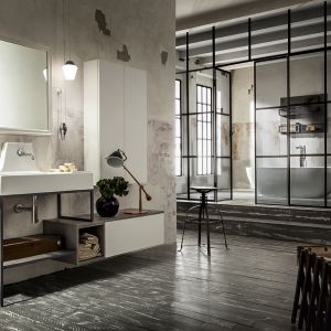 Aranżacja łazienki w stylu industrialnym zaproponowana przez firmę Cerasa, z kolekcją mebli Urban Play. Białe matowe fronty zestawiono z metalowym szkieletem. Ścianę pokrywa obdrapany tynk, a na podłodze widnieje postarzana deska. Charakterystycznym akcentem jest laboratoryjny stołek. Fot. Cerana