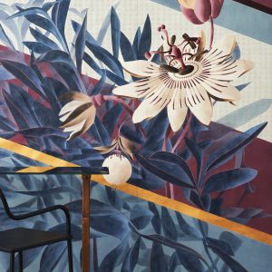 „Passiflora”, czyli kwiat męczennicy z owocami, jest niekwestionowanym bohaterem tej tapety. Ponadwymiarowy design łączy się tu z bujną paprocią poprzez naprzemienne nakładanie się i przenikanie elementów poszczególnych planów.
Inspiracją dla tła jest świat tekstyliów, gdzie geometryczne mikro wzory występują naprzemiennie z szerokimi ukośnymi paskami. Na jej wyjątkowy wygląd wpływają dodatkowo ręcznie haftowane detale, które nadają kwiatom aspekt pełnej trójwymiarowości. Fot. Mood-Design
