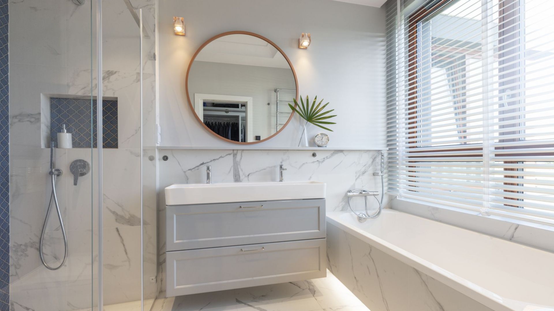 W jasnej łazience głównym motywem aranżacyjnym są płytki imitujące marmur. Ciemniejszą kolorystykę zastosowano w strefie prysznica. Projekt: Decoroom. Fot. Pion Poziom